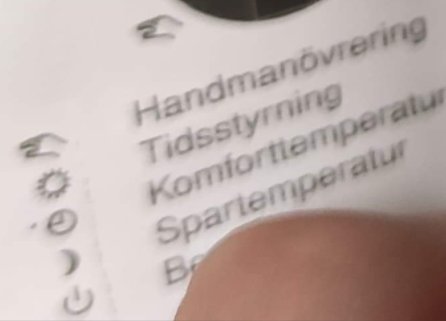 Närbild på en termostat med texten 'Handmanövering, Tidsstyrning, Komforttemperatur, Spartemperatur' och symboler, otydligt finger i förgrunden.