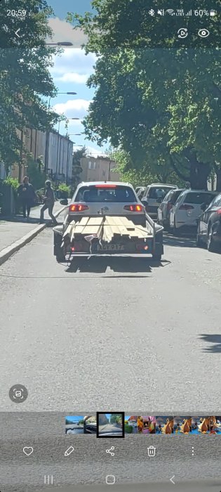 Bil som drar ett släp med virke på en stadsgata, människor i bakgrunden.