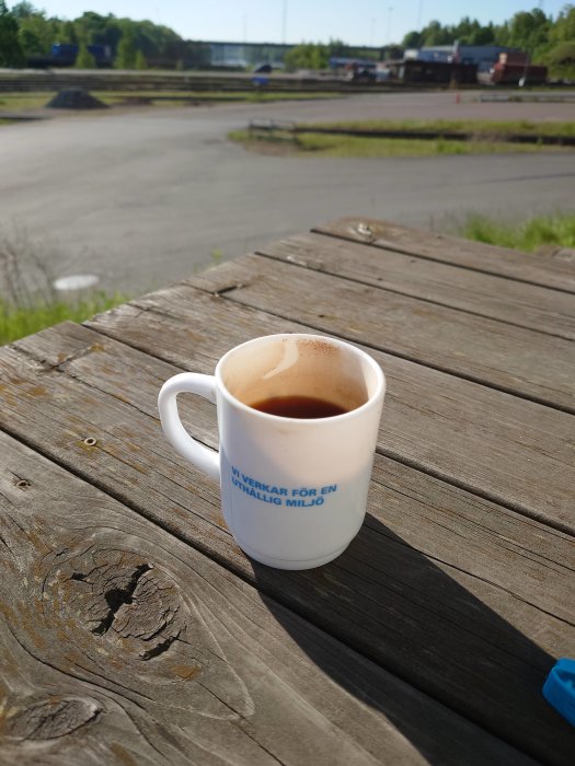 Kaffekopp på träbord med texten "Vi verkar för en uthållig miljö" utomhus på en solig dag.