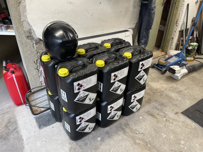 Storpack med svarta dunkar märkta med farosymboler staplade i ett garage med bygghjälm och brandsläckare.