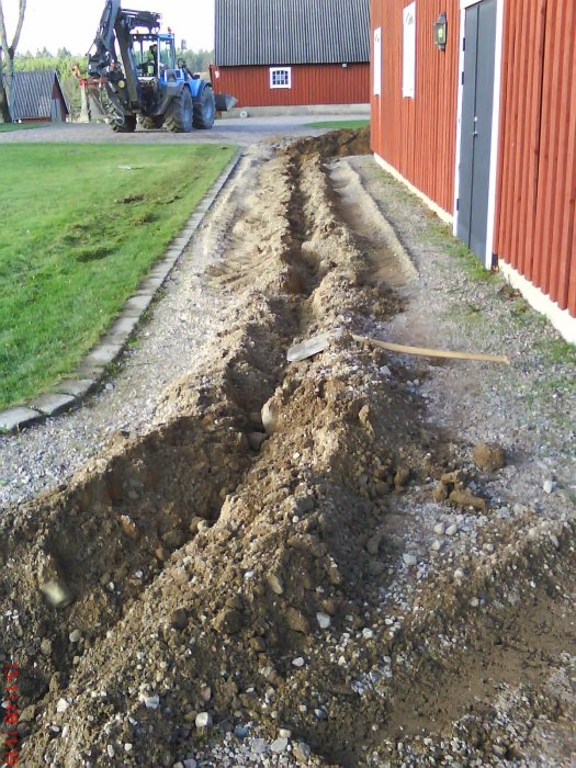 Grävmaskin vid nygrävd ränna för kabelläggning längs en grusväg med röd ladugård i bakgrunden.