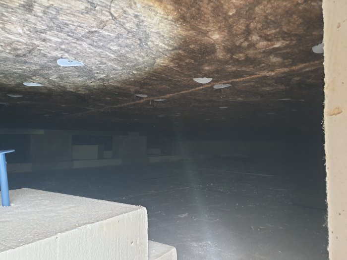 Vy under krypgrund med spruckna spikbrickor som fäster isolering mot golvbjälkar.