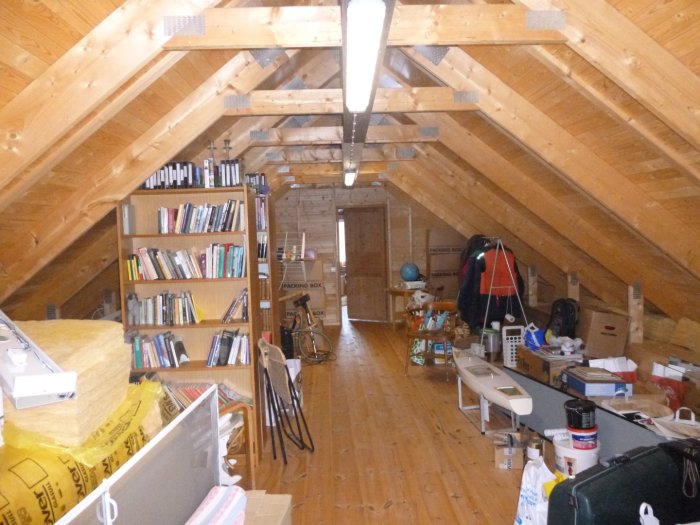 Vind med träbjälkar, förrådshyllor, sängar och bord under snedtak.