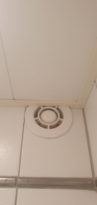 En vit fläkt insatt i ett ventilationsrör på väggen, med kakel och en hörnlist runt omkring.