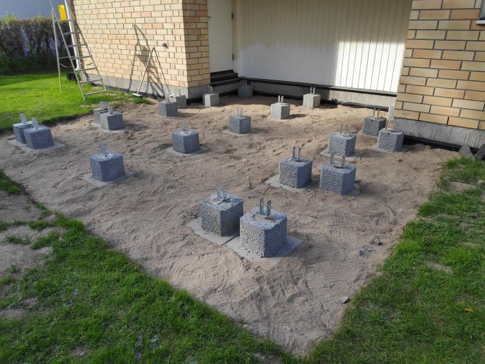 Grund med stolpskor fastgjutna i betongblock jämnt fördelade på marken vid husvägg.