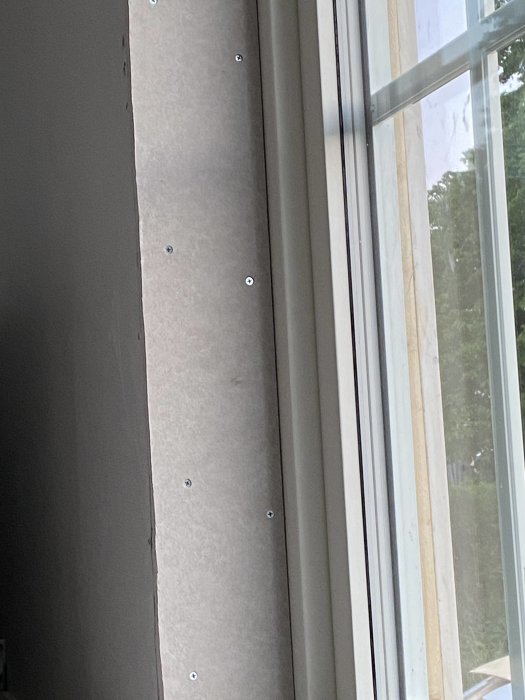 En grå byggskiva monterad vertikalt bredvid ett fönster, fäst med skruvar vid sidan i ett rum under renovering.