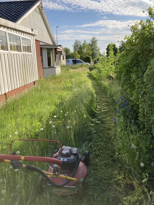 Gräsklippare framför nyklippt stig bland högt gräs bredvid ett hus, känsla av prestation.