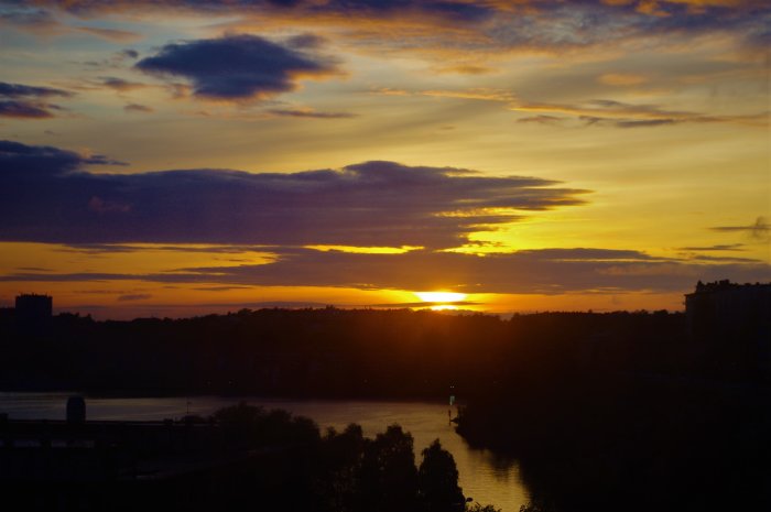 Solnedgång över ett landskap med flod och silhuetter av byggnader mot en färgstark himmel.