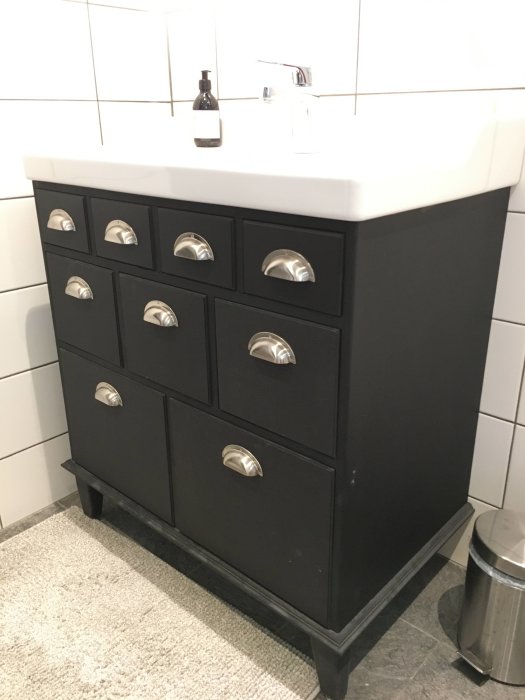 Svart badrumskommod med handfat och silverfärgad blandare mot vita kakelväggar.