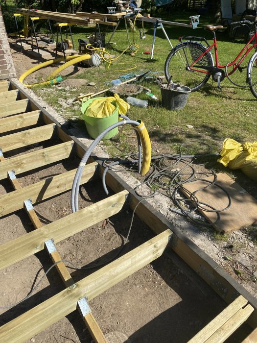 Byggarbetsplats för trädäck med träbalkar och balkskor, verktyg och cykel i bakgrunden.