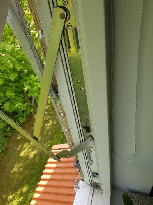 Fönstrets gångjärn och undre karm syns, med tydligt slitage och skador nära fönsterhandtaget.