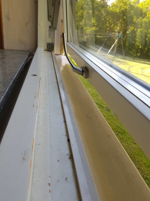 Slitet fönsterkarm nedre del utan synlig plåt, smuts och skador synliga, med utsikt över trädgården.