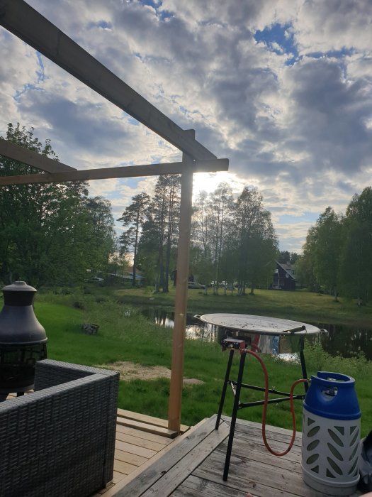 Kvällsutsikt från en veranda med en oavslutad träkonstruktion, bord, stolar och en utomhuskamin, med träd och en sjö i bakgrunden.