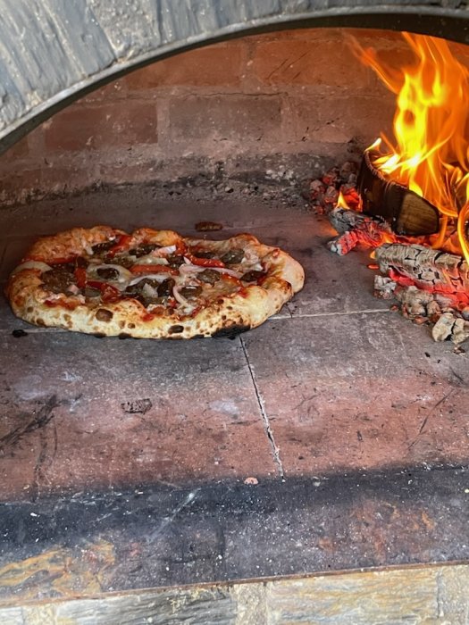 Pizza som gräddas i en vedeldad ugn med synliga lågor till höger.