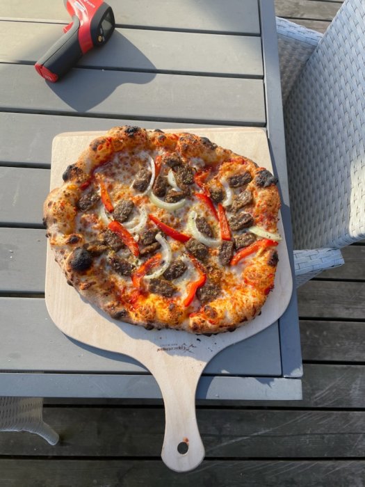 Nybakad pizza med korv och paprika på en träspade bredvid en gasolbrännare på ett utomhusbord.