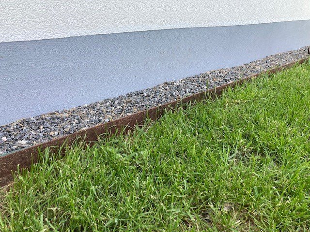 Rabattkant av cortenstål mellan gräsmatta och makadambelagd kant vid en husvägg.