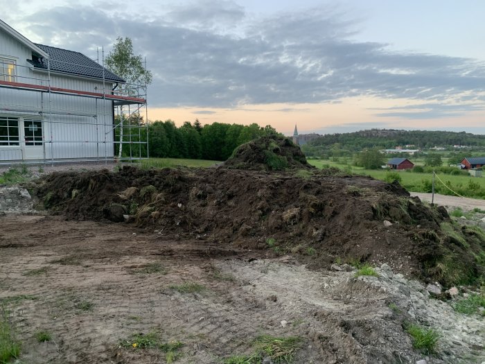 En hög med grävd jord framför ett hus under konstruktion med ställningar och skymning i bakgrunden.