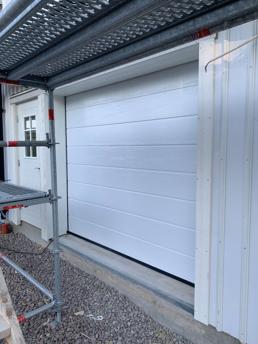 Nyinstallerad vit garageport på ett hus under byggnation, omgiven av byggställning och grusgång.