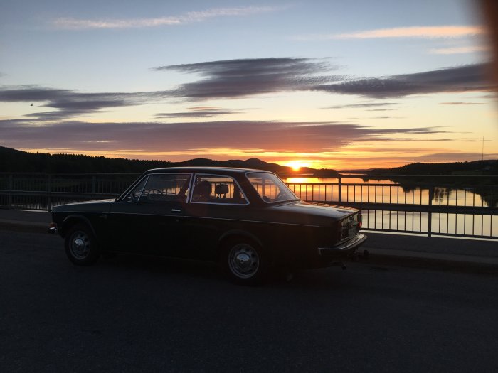 Cypressgrön veteranvolvo 142 från 1972 parkerad vid en bro i solnedgången med spegelblank flod i bakgrunden.