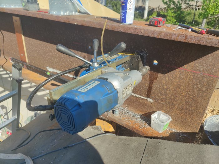 Borrmaskin som kärnborrar i en stor stålbalk, med borrspån och verktyg synliga runtomkring.