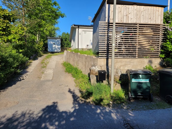 En sliten garageuppfart med ojämn mark och sprucket underlag omgiven av grönska och en trästaket.