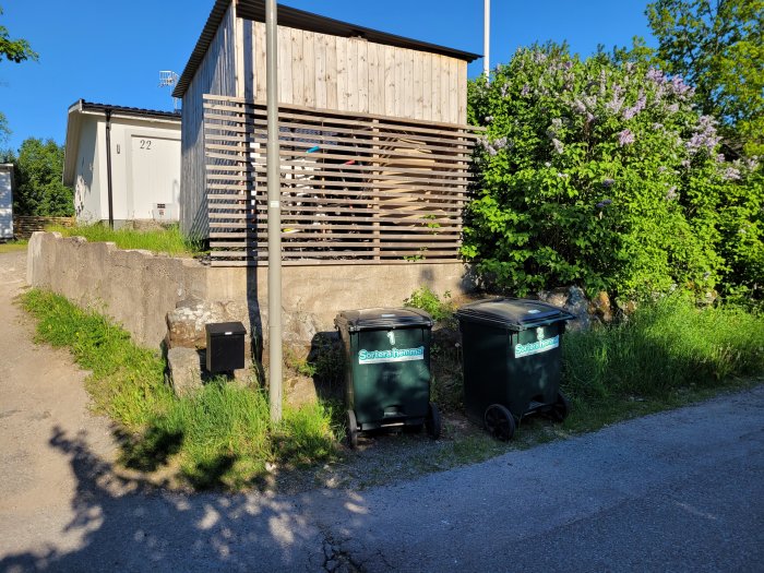 Uppfart vid ett hus med grusväg, stenmur och sopkärl, saknar stenläggning.