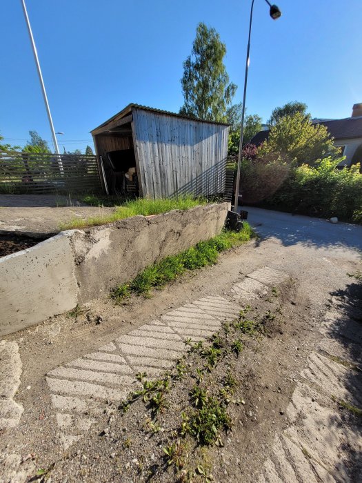 Ojämn garageuppfart med sliten marksten och grus framför en gammal, plåtbeklädd byggnad på en solig dag.