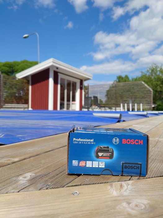 Bosch-batteri på trägolv utomhus med rödvit stuga och blå himmel i bakgrunden.