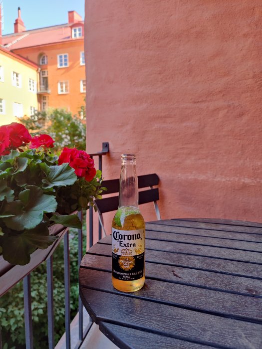 Flaska Corona Extra på ett svart bord ute på solig balkong med röda pelargoner och färgglada hus i bakgrunden.