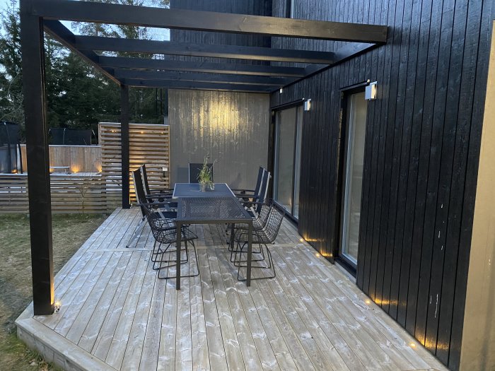 Modern uteplats med trädäck, svart pergola, matbord med stolar och integrerad belysning intill en svart träfasad.