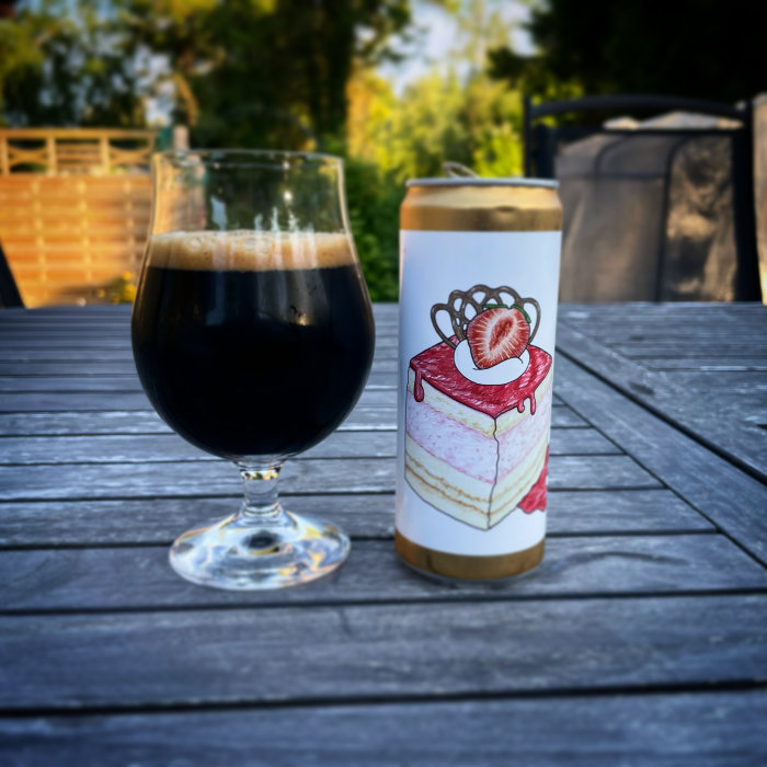 Mörk öl i glas bredvid ölburk med tårtdesign utomhus på träbord.