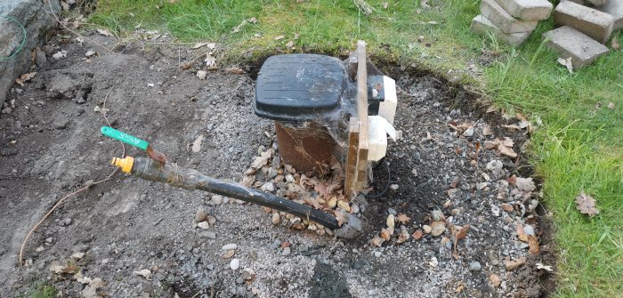 En öppen brunn med svart lock synlig i en trädgård, med slang och elpanel, omringad av grus och löv.