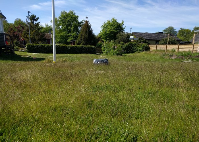 En robotgräsklippare "Tuva" som arbetar på en gräsmatta med trädgård och hus i bakgrunden.