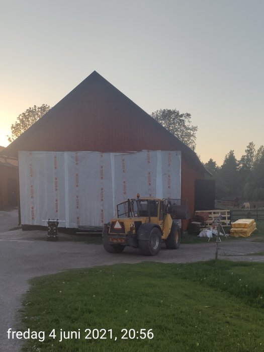 Gaveln på en röd lada skyddad med byggplast och en gaffeltruck i förgrunden, kvällshimmel i bakgrunden.