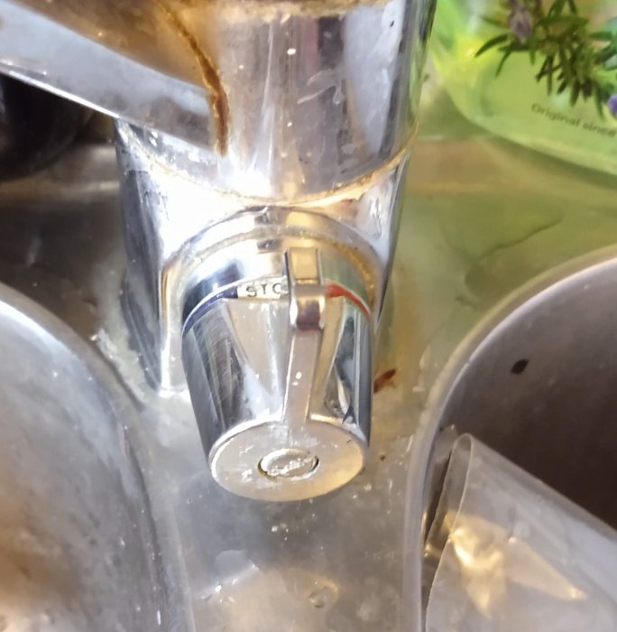 Närkymd bild av en kromad köksblandare med tydlig kalkavlagring och en markerad avstängningsknapp för diskmaskin.