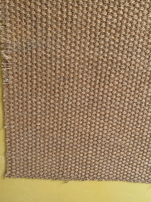 Del av en borttagen sisalmatta med synlig vävd textur och gula limrester på ett underliggande golv.