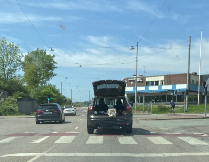 Bil med öppen baklucka full med lösa föremål som bollar och burkar på en trafikerad väg.