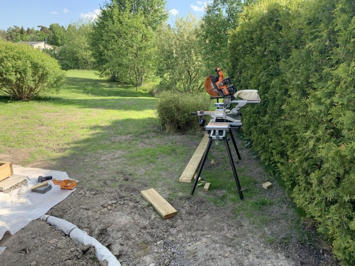 Byggplats utomhus med kap-/gersåg på stativ, träreglar och arbetsverktyg, i en trädgård med vacker grönska.