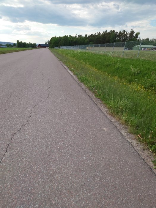 Asfalterad landsväg med sprickor, gräsbeväxt vägren och staket längs med vägen under molnig himmel.