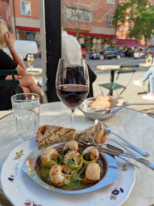 Tallrik med sniglar i vitlökssmör på uteservering med vin och bröd, i stadsmiljö.