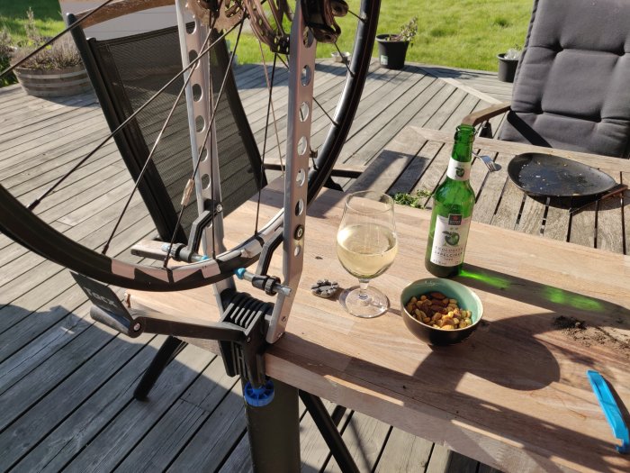 Cykel i riktställ på en träterrass, ciderflaska, glas och skål med nötter bredvid verktyg och cykeldelar i solljus.