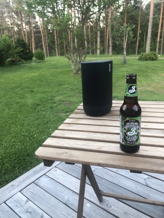 En bärbar högtalare och en flaska Brooklyn Lager på ett träbord utomhus.