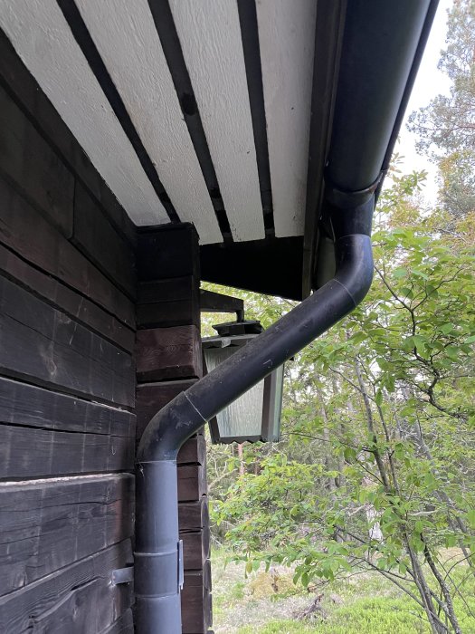 Hörn av hus med mörkbrun brädfasad och vitmålade bjälkar under ett tak, med ett stuprör och grönska i bakgrunden.