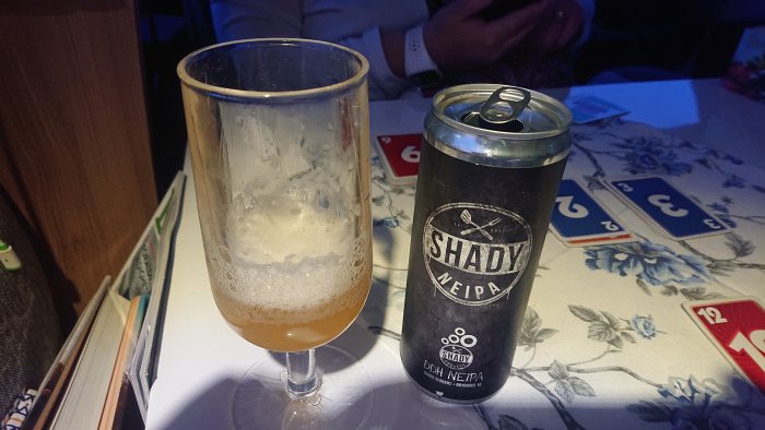 Halvtomt ölglas bredvid en burk Shady NEIPA från Brygghus 19 på ett blommigt bord.