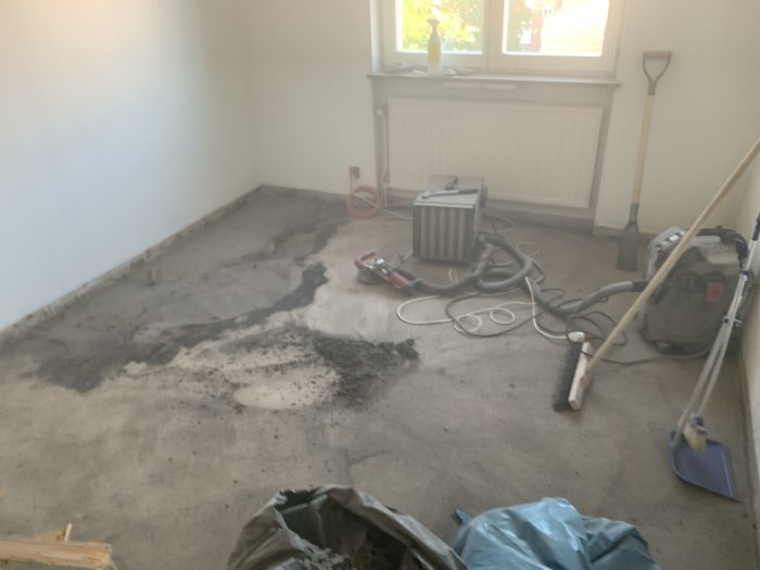 Ett rum under renovering med slipmaskin, dammsugare och städredskap på ett delvis slipat betonggolv.