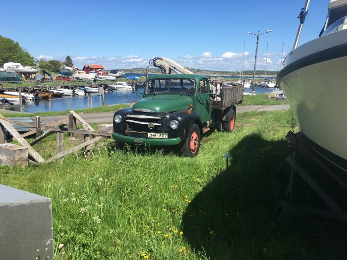 Gammal grön lastbil med flak parkerad på gräs nära båthamn med båtar och bryggor i bakgrunden.