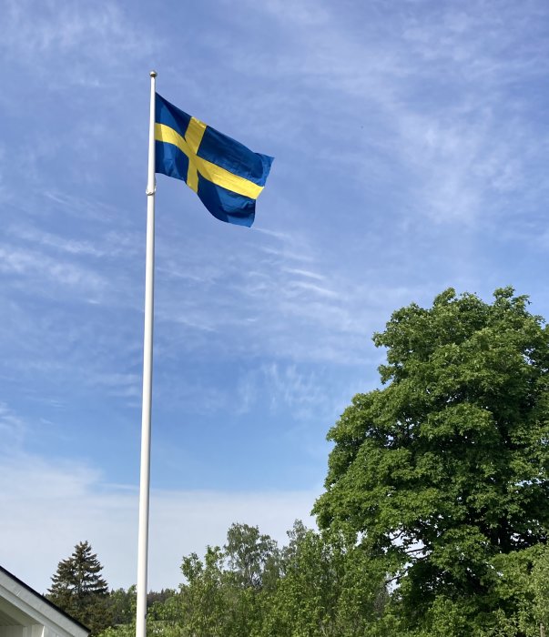 Svenska flaggan vajar mot en blå himmel ovanför gröna träd.