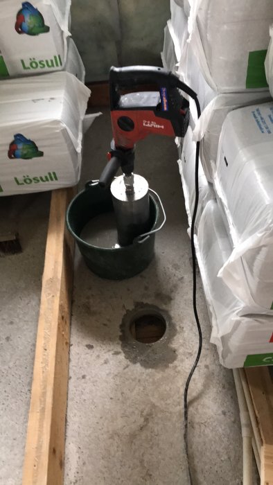 Hilti borr med krona i hink med vatten bredvid borrat 130 mm hål i betonggolv, omgivet av byggmaterial.