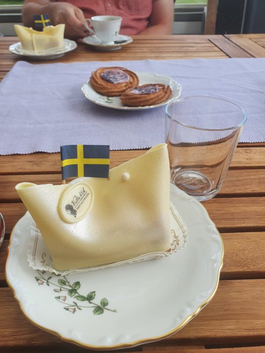 Fikadags med svensk prinsesstårta och kanelbullar på ett träbord, en person i bakgrunden dricker kaffe.