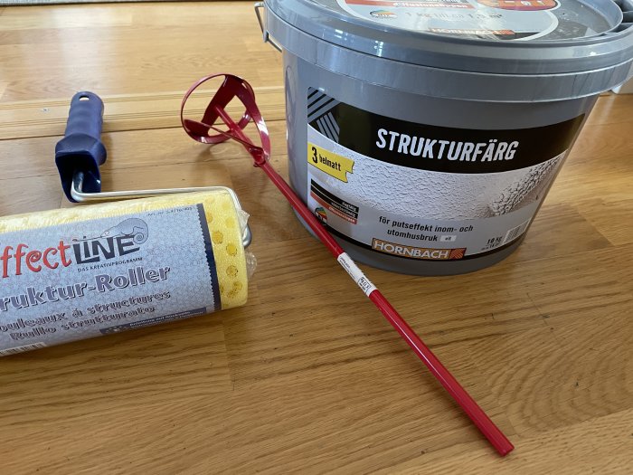 Strukturfärg i hink, strukturroller och rörverktyg för borrmaskin på trägolv.